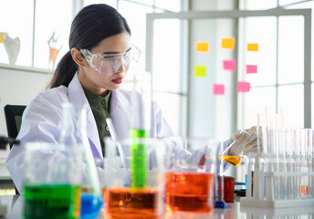 Asian woman as chemistry expert pouring experiment liquid from beaker into glass tube with safety protection of goggles and glove at scientific lab