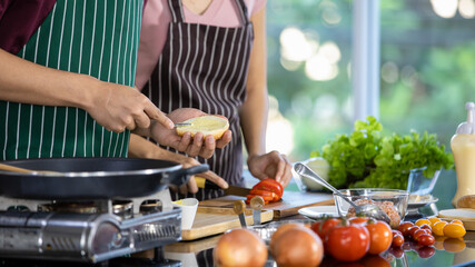 Asian chef cooking tasty food by spreading butter on toasted bread and slicing tomato into pieces on cutting board beside frying pan on gas stove, fresh vegetable for dining party.