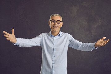 Portrait of cheerful senior man in eyeglasses spreading his arms wide open standing against grey studio background. Smiling retired aged teacher happy to hug his students and give them a warm welcome