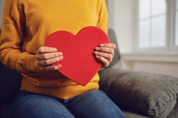 Valentine's day. Red heart in the hands of a girl in casual clothes sitting on the couch in the room.