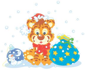 Cute little tiger in a red and white Santa hat sitting on snow with a beautiful gift bag and a Christmas toy ball, vector cartoon illustration isolated on a white background