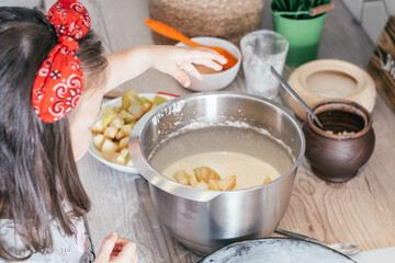 Little dark-haired girl 3 years old in red headband bakes apple pie in kitchen. Child adds apples to dough. Kid cooking