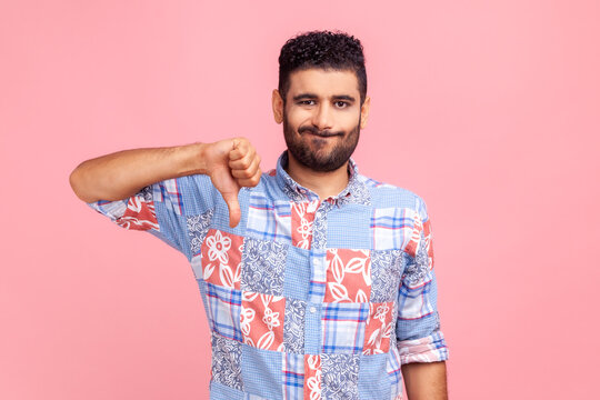 Portrait of young adult man in blue shirt criticizing bad quality with thumbs down displeased grimace, showing dislike gesture, expressing disapproval. Indoor studio shot isolated on pink background.
