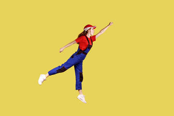 Fototapeta na wymiar Side view portrait of woman worker flying to do her work like superhero, fast and high quality service, wearing work uniform and red cap. Indoor studio shot isolated on yellow background.