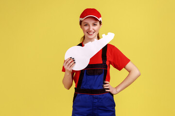 Smiling beautiful worker woman looking at camera with positive emotions, holding big paper key, estate agency, wearing work uniform and red cap. Indoor studio shot isolated on yellow background.