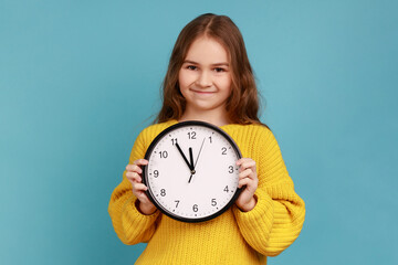 Portrait of little girl holding big wall clock in hands, looking at camera with happy expression,...