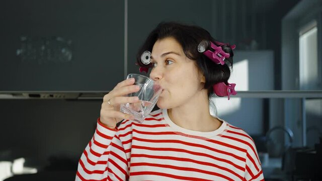 Woman with hair curlers wearing striped shirt drinking water in kitchen, black cupboards on background. Portrait of housewife in the morning. Concept of healthy lifestyle