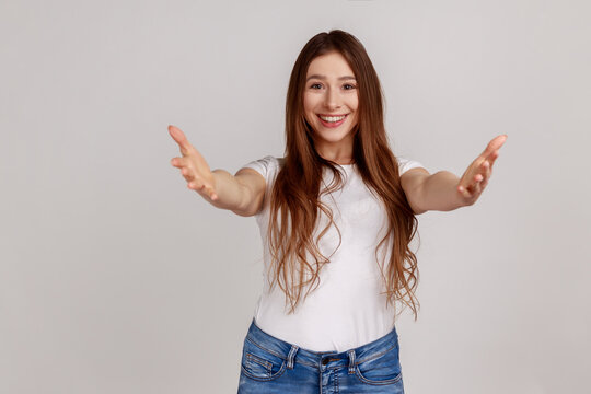 Portrait of friendly woman giving free hugs with outstretched hands, welcoming inviting to embrace, support and care, wearing white T-shirt. Indoor studio shot isolated on gray background.
