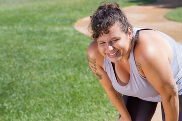 Naklejka premium Portrait of woman with curly hair doing sports on sunny day. Chubby woman in grey top with tattoos looking sideways. Sport, body positive concept