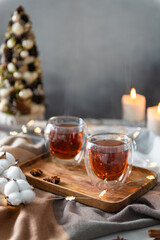 Cozy winter composition with hot tea in glass cup with steam. Two cups of black tea with steam on wooden tray on plaid with candles and Christmas tree. Copy space. Christmas menu, winter tea party