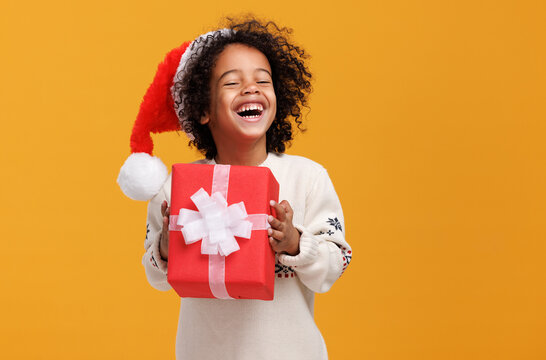 Happy little afro american curly boy in santa hat and knitted sweater hugging big Christmas gift box tightly