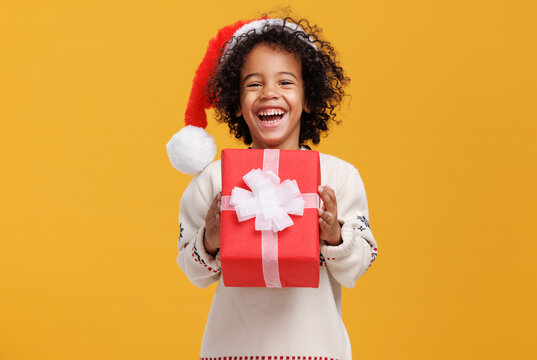 Happy little afro american curly boy in santa hat and knitted sweater hugging big Christmas gift box tightly