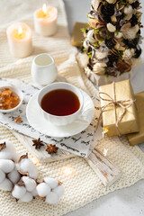 Winter hot tea in a cup in cozy composition. A Cup of black tea on wooden tray on knitted plaid with candles, gifts and Christmas tree. Side view. Christmas menu, greeting card, poster, winter tea 