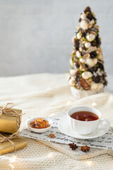 Winter cozy background with cup of tea on wooden tray, gifts, candles, branch of cotton and christmas tree. Winter menu, tea party, poster, greeting card. Product place, copy space, hygge