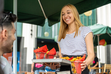 Young blonde waitress is serving French fries and beer to customers at the table, catering business and restaurant workers