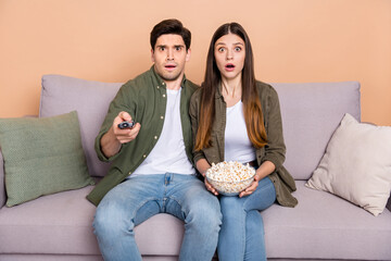 Photo of frightened spouses sit couch use remote control hold pop corn bowl watch series isolated...