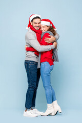 Full length portrait of happy Caucasian couple in Christmas outfits huging each other in light blue...
