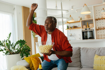 African pensioner sitting at his cozy home, watching television and eating popcorn. He is enjoying...