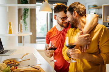 Happy hipster gay couple in love hugging while standing in the kitchen. They are cooking dinner together at home and drinking wine - obrazy, fototapety, plakaty