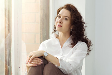 Young beautiful woman with curly hair in a white shirt sits on the windowsill. The concept of melancholy, unrequited love, self-acceptance, spiritual growth. Place for text