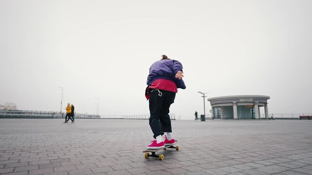 Skating youth hobby concept. Young good looking skate boy in a purple jacket and red shoes is practicing longboarding at the skate in an empty city square