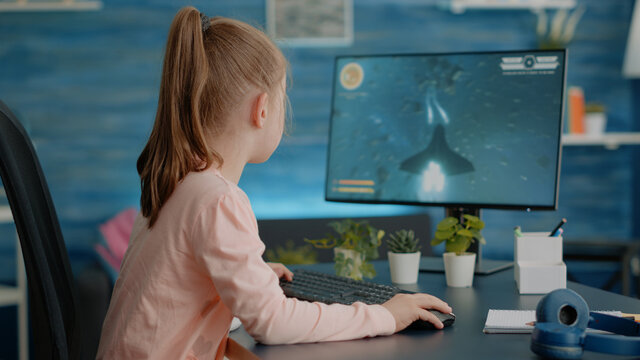 Schoolgirl using computer to play video games for entertainment at home. Child playing action shooting game with monitor and keyboard, enjoying fun activity after doing homework.