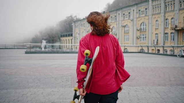Portrait of a young teenager with a skateboard under his arm. A long-haired trendy man wearing a red shirt walks through the town square. Hipster portrait with longboard, back view