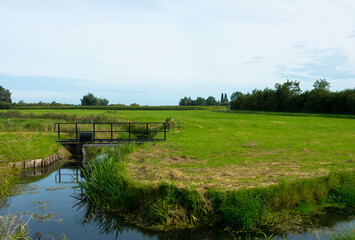 Dutch countryside landscape, Typical polder and water land, Green meadow with footbridge on the blue sky. Small canal or ditch on the field, Friesland Netherlands