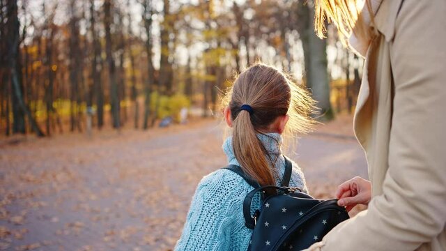 Young mom putting plastic container into backpack of little daughter, zipping it. Hugging, smiling and walking away together. Autumn, park. Close up