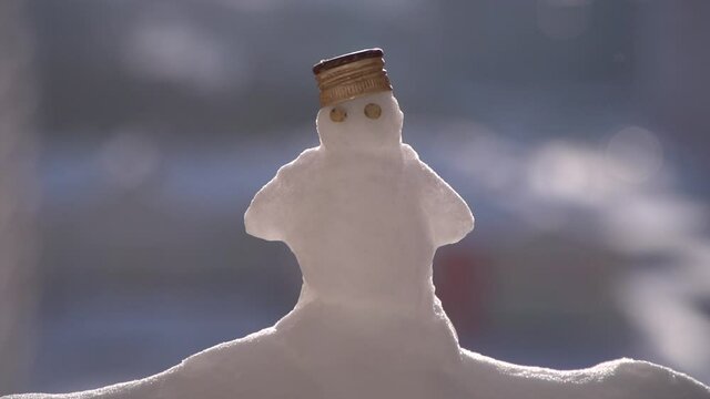 Little snowman with pea eyes and bottle metal cap on head standing on snowdrift when snowing and sunny. Snowman is in the center of the frame, background is out of focus