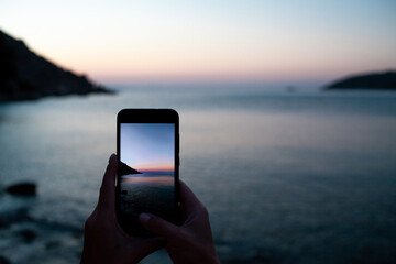 woman hands holding mobile phone at sunrise over sea bay. Taking photo of sunset over rocks in water by mobile phone. Selective focus on smartphone