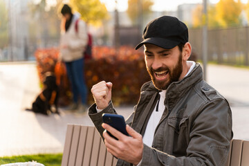 Portrait of happy guy gesturing and rejoicing while locating at the bench at the street with his mobile phone. Brunette man browsing internet at the fresh air