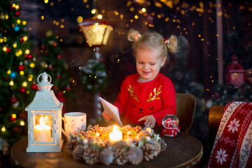 Fototapeta na wymiar Cute little blond child, curly girl with two ponytails, sitting on a table with christmas decoration, writing letter and playing with toys