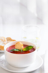 Tomato soup in white bowl decorated with bread crumbs, fresh basil  and herbs on the restaurant table with water glass and portion of bread .
