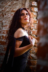 Fototapeta premium A brunette girl with long hair in an evening dress in a vintage Gothic castle. A woman who looks like an evil witch or sorceress in an abandoned old church. Female Model posing in Halloween