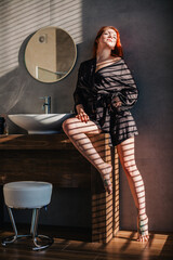 A young, slender, red-haired girl in a black bathroom robe stands in the sunlight through blinds