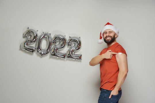 A happy man with a beard in a Santa hat is showing a shoulder with a plaster after vaccination against Coronavirus (Covid-19) near foiled silver balloons in the shape of 2022.