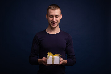 Smiling young man in a blue sweater posing on a blue background, holding a gift in his hands