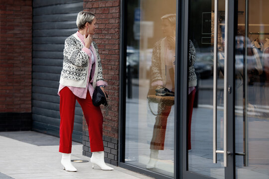 Fashionably dressed woman shopping, looking at reflection in mirror of shop window outside. Adult model with short hair wearing sweater, red pants. Candid photos of people in public places