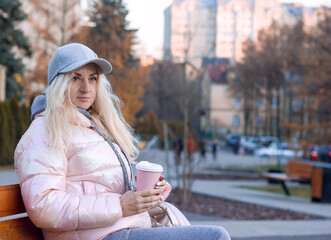 Young attractive woman in a warm pink jacket and a cap sits on a bench on a city street and holds in her hands a disposable glass with a hot drink coffee tea