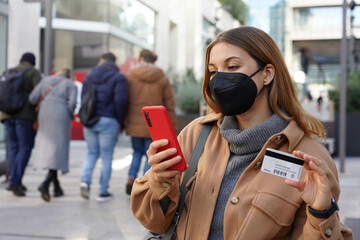 Fototapeta premium Credit card payment outdoors. Beautiful young woman with black protective mask FFP2 KN95 enters her credit card number on smart phone for shopping online in city street.