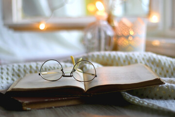 Open book and reading glasses. Candles and fairy lights in the background. Selective focus.