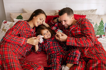 Happy young family in red pajamas playing on the bed on Christmas Eve in their apartment with New Year's decor. Dad, mom and daughter are lying on the bed and having fun. Family celebrating christmas