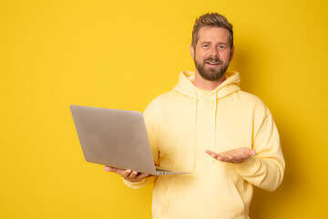 Portrait of a happy young man holding laptop computer isolated on yellow background.