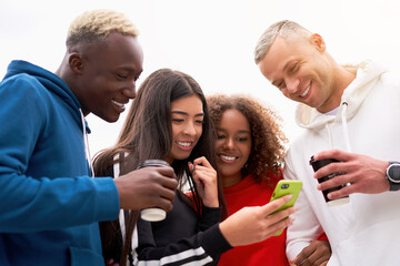 Young people, students of different nationalities, smile, hold coffee in their hand and look into one smartphone. Spend time together with friends. Two men and two women. White background.