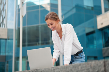 Blonde business woman in a white blouse uses a laptop to work while outside, working outdoors. In the background is a business center.