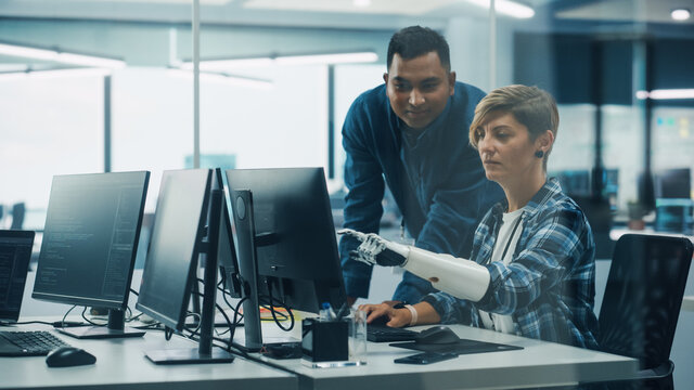 Teamwork In Diverse Inclusive Office: Project Manager Talks with Woman with Disability with Prosthetic Arm Work on Desktop Computer. Professionals Software Engineers Create e-Commerce App Solution - Powered by Adobe
