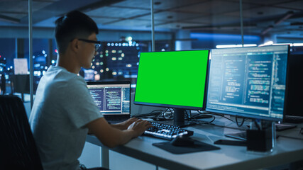 Night Office: Young Japanese Man in Working on Green Screen Chroma Key Desktop Computer. Programmer Typing Code, Creating Modern Software, e-Commerce App Design. Over Shoulder