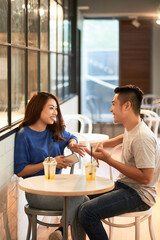 Smiling young Asian couple in casual outfits sitting at small table with drinks and chatting together in cafe