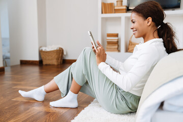Young black woman using cellphone while sitting on floor at home